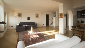 Amplia sala de estar con un sofá grande, una mesa de centro y una alfombra estampada. Se ve una escalera que sube a un nivel superior, y un comedor con una mesa y sillas.