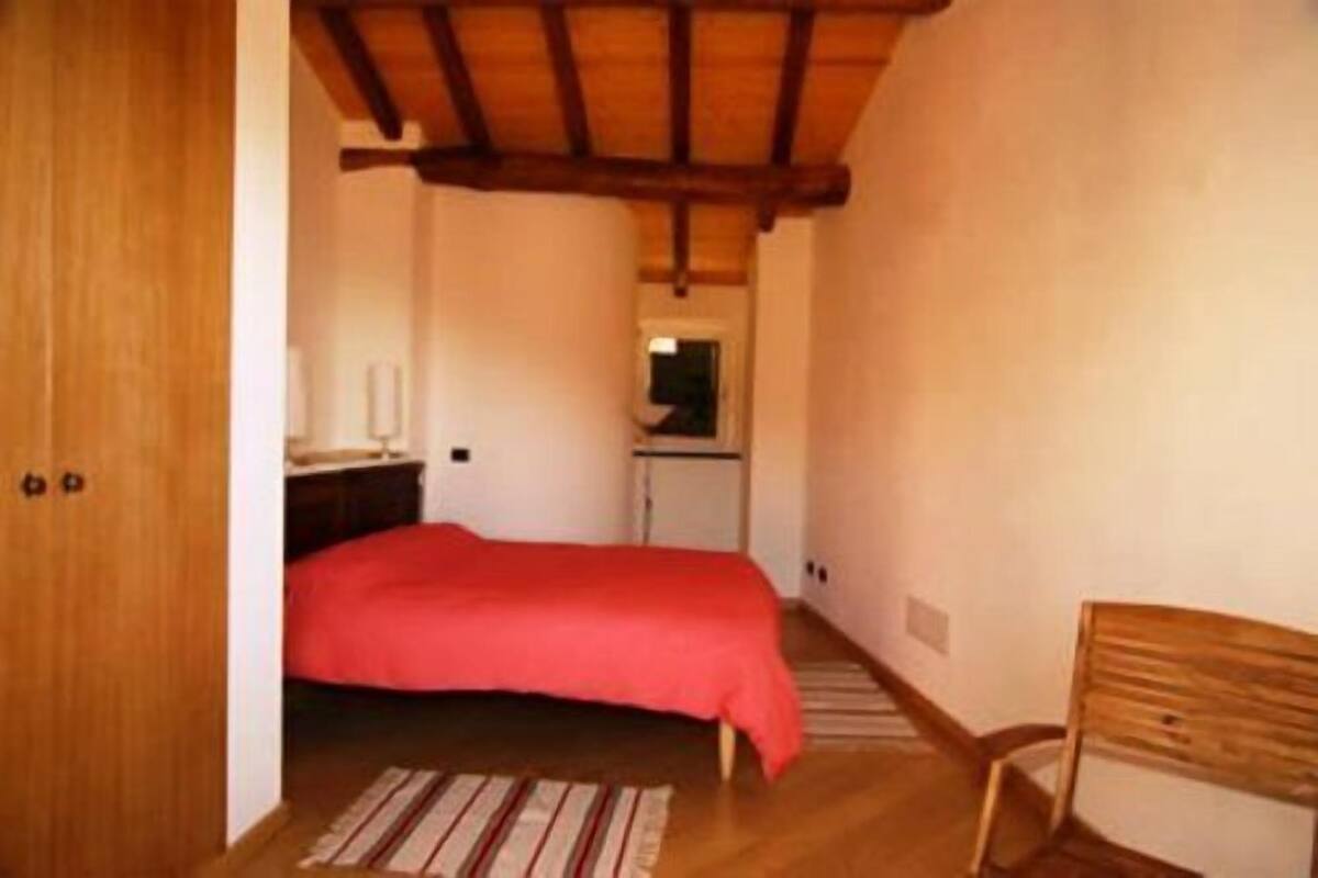 A bedroom with a wooden ceiling, a bed with a red bedspread, a wooden chair, and a small kitchenette in the background.