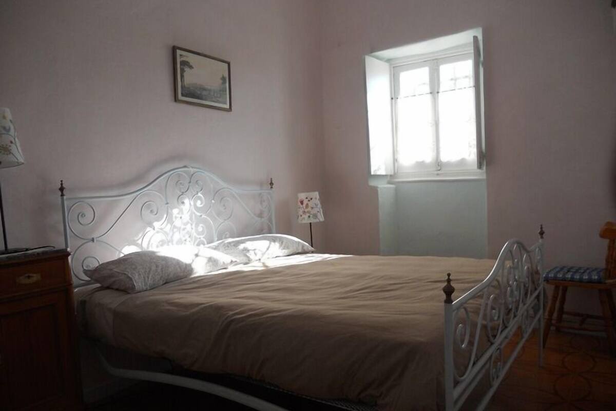 A bedroom with a bed, a nightstand, a chair, a window, and a wall-mounted picture.