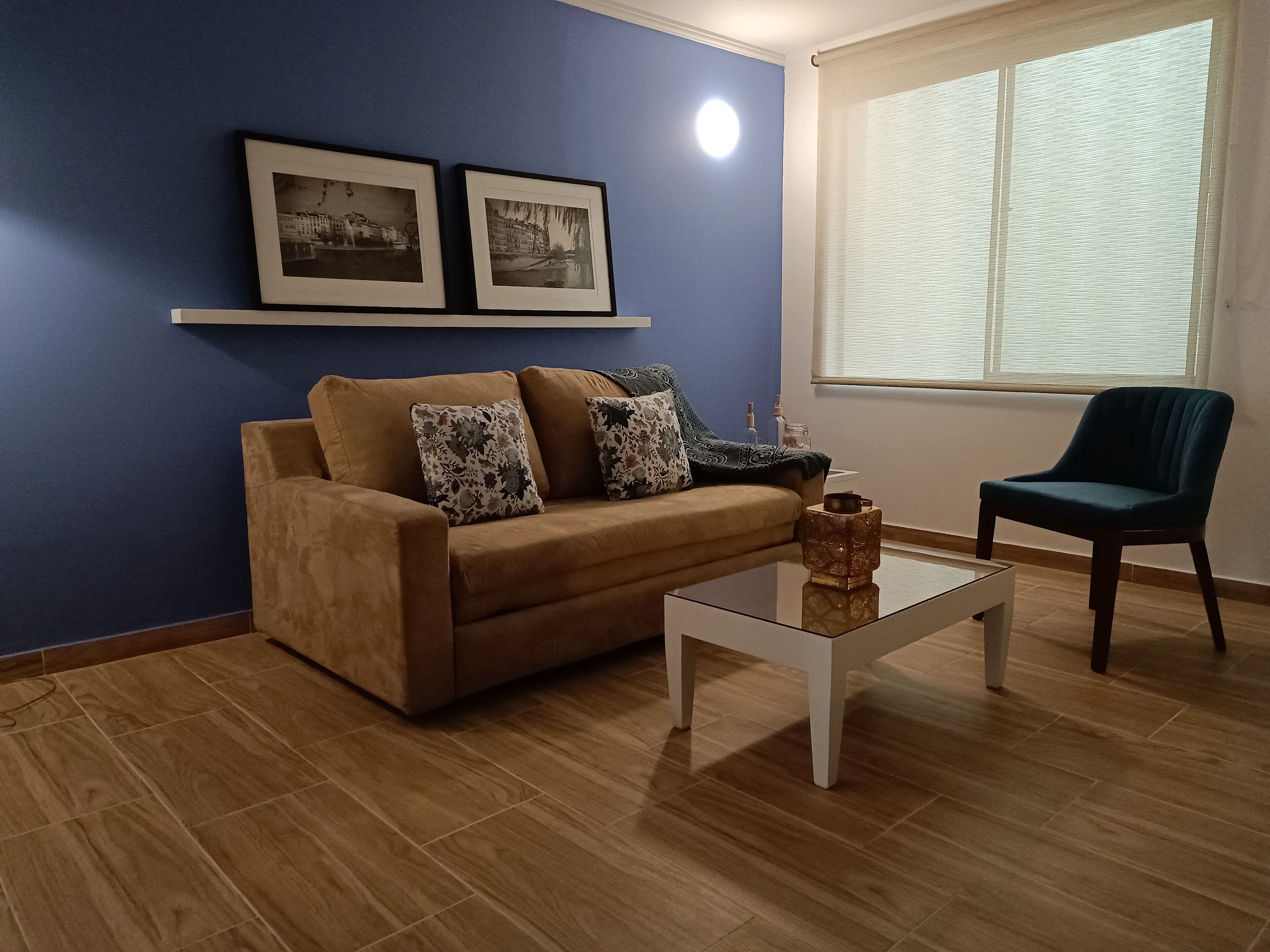 A living room with a brown sofa, a glass coffee table, a blue armchair, and two framed pictures on the wall.