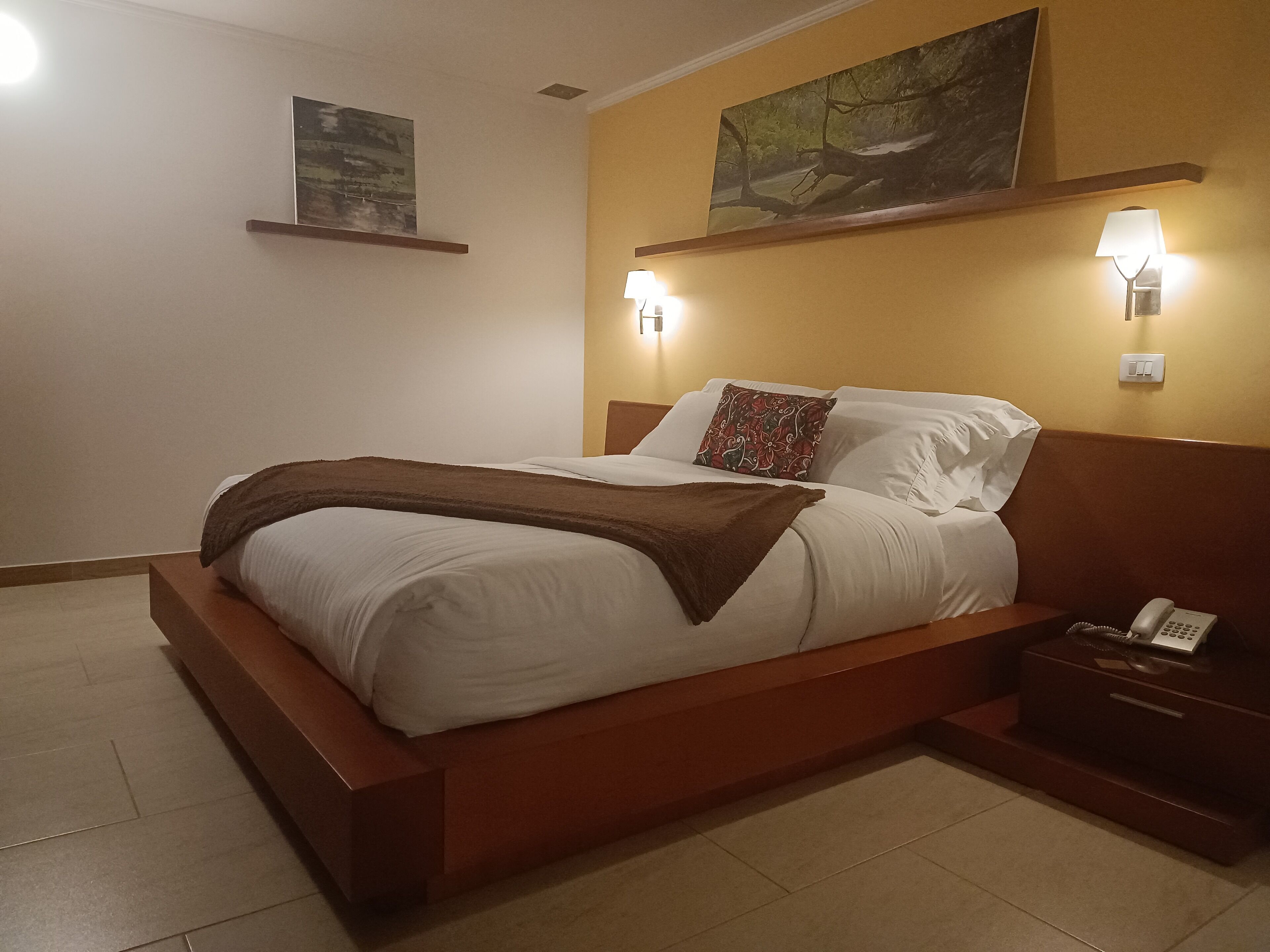 Un letto rifatto con cura, una coperta marrone, un telefono sul comodino e due lampade a parete.