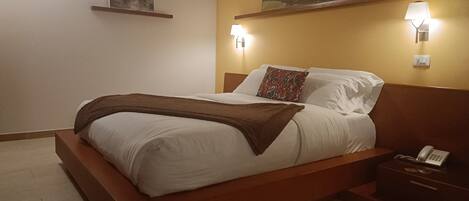 Una cama bien hecha con una manta marrón, un teléfono en la mesita de noche y dos lámparas fijadas a la pared.