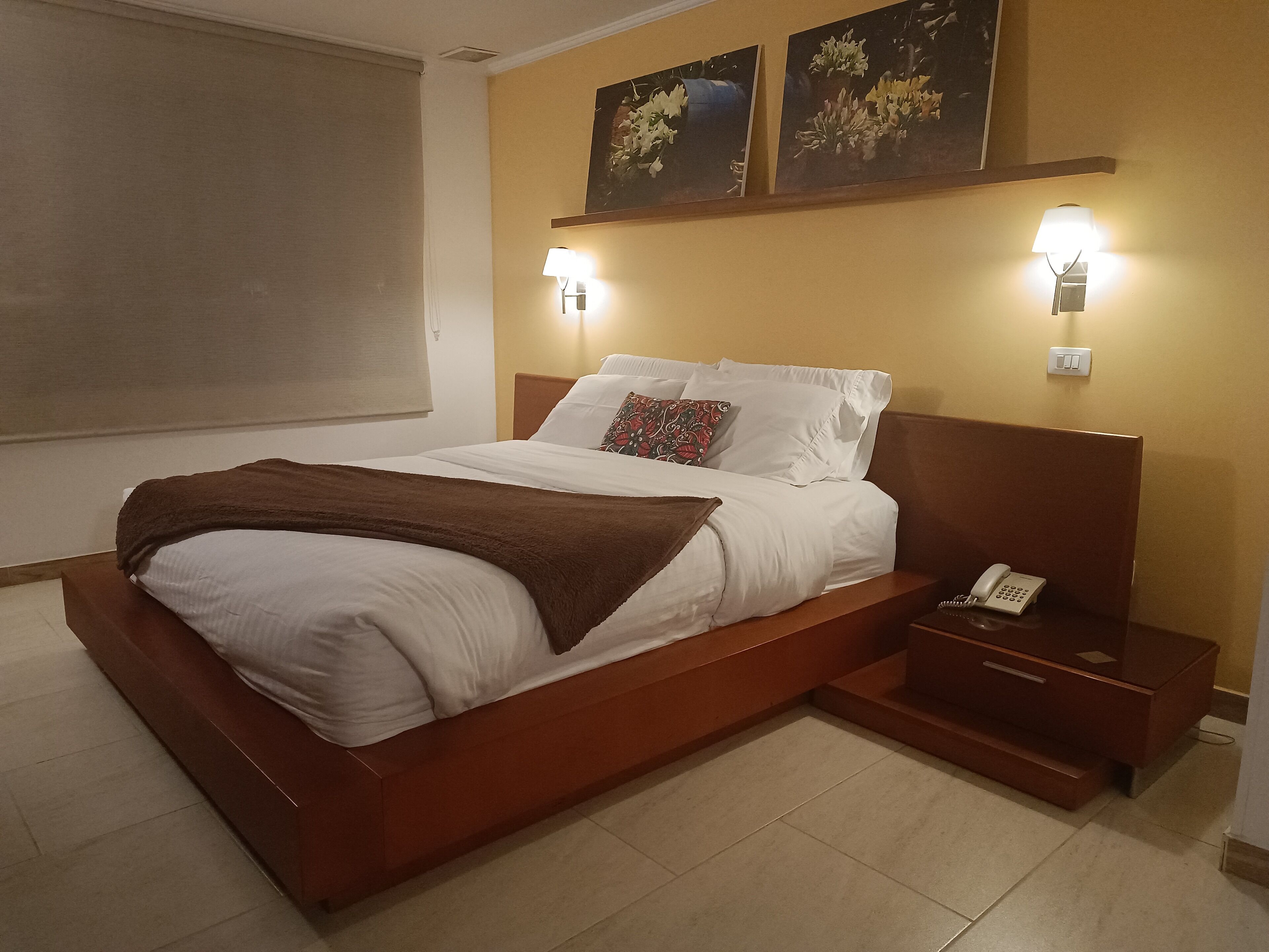 Una cama bien tendida con sábanas blancas y un plaid marrón, un teléfono en la mesita de noche y dos lámparas de pared.