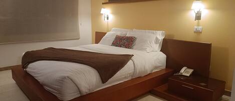 Una cama bien tendida con sábanas blancas y un plaid marrón, un teléfono en la mesita de noche y dos lámparas de pared.