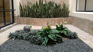 Un patio con pared de ladrillo, zona de grava negra y diversas plantas.