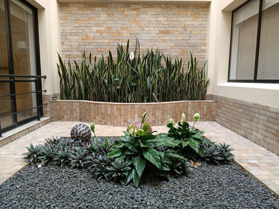 Un patio con pared de ladrillo, zona de grava negra y diversas plantas.