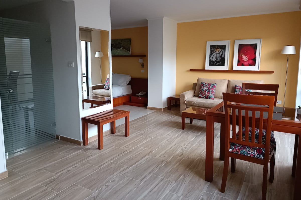 Une chambre d’hôtel moderne avec un lit, un canapé, une table basse en bois et une chaise. Il y a un miroir au mur, une lampe et un cadre photo.
