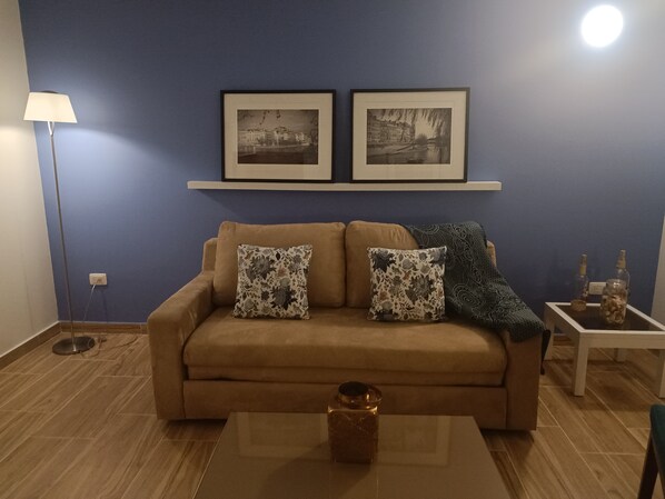 Una sala de estar con un sofá color beige, dos cojines estampados, una mesa de centro de vidrio, una lámpara de pie y dos cuadros en la pared.