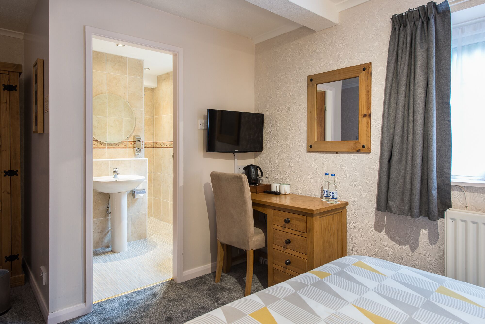 A hotel room with a bed, a desk with a chair, a television, a bathroom with a sink and mirror, and a window with curtains.