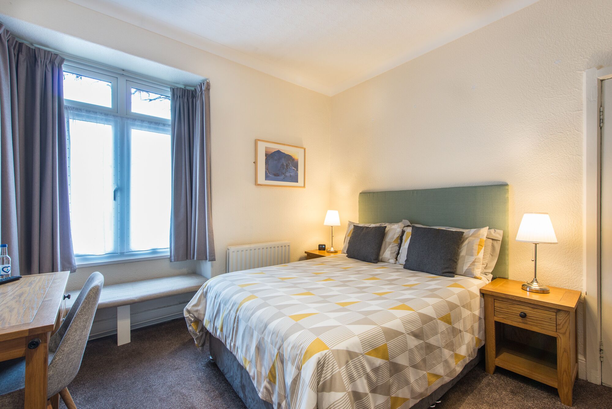 A hotel room with a bed, two bedside tables, a desk with a chair, a window with curtains, and a radiator.
