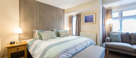 A hotel room with a bed, bedside table, lamp, window with curtains, and a sofa.