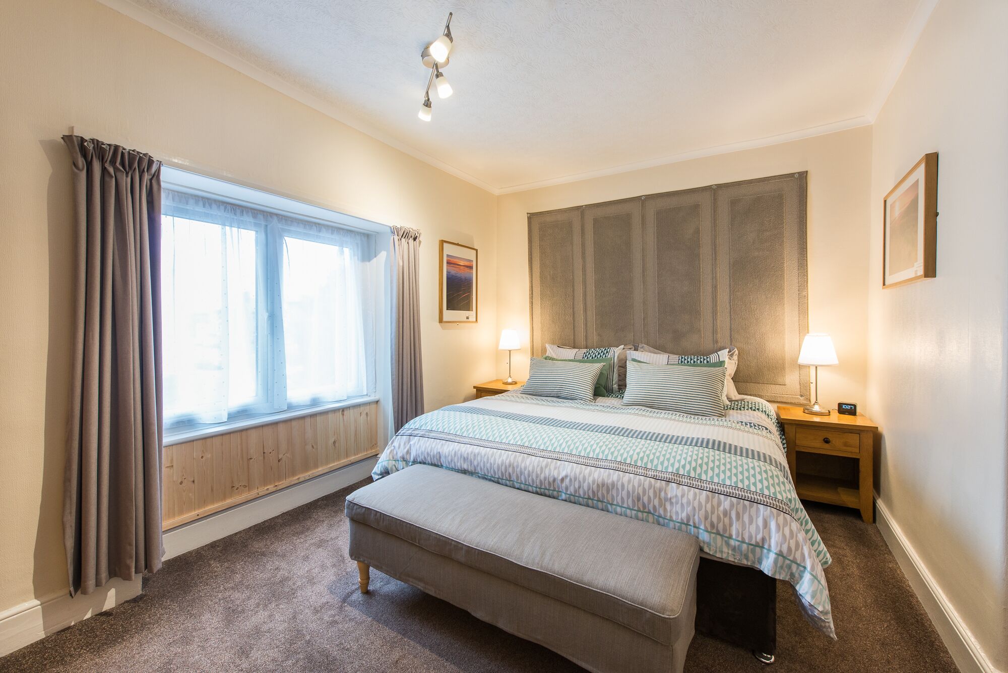 A bedroom with a large bed, two bedside tables, a nightstand, a window with curtains, and a ceiling light.