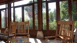 Eine überdachte Veranda mit Holzstühlen und einem Tisch, großen Fenstern und Blick auf Bäume.