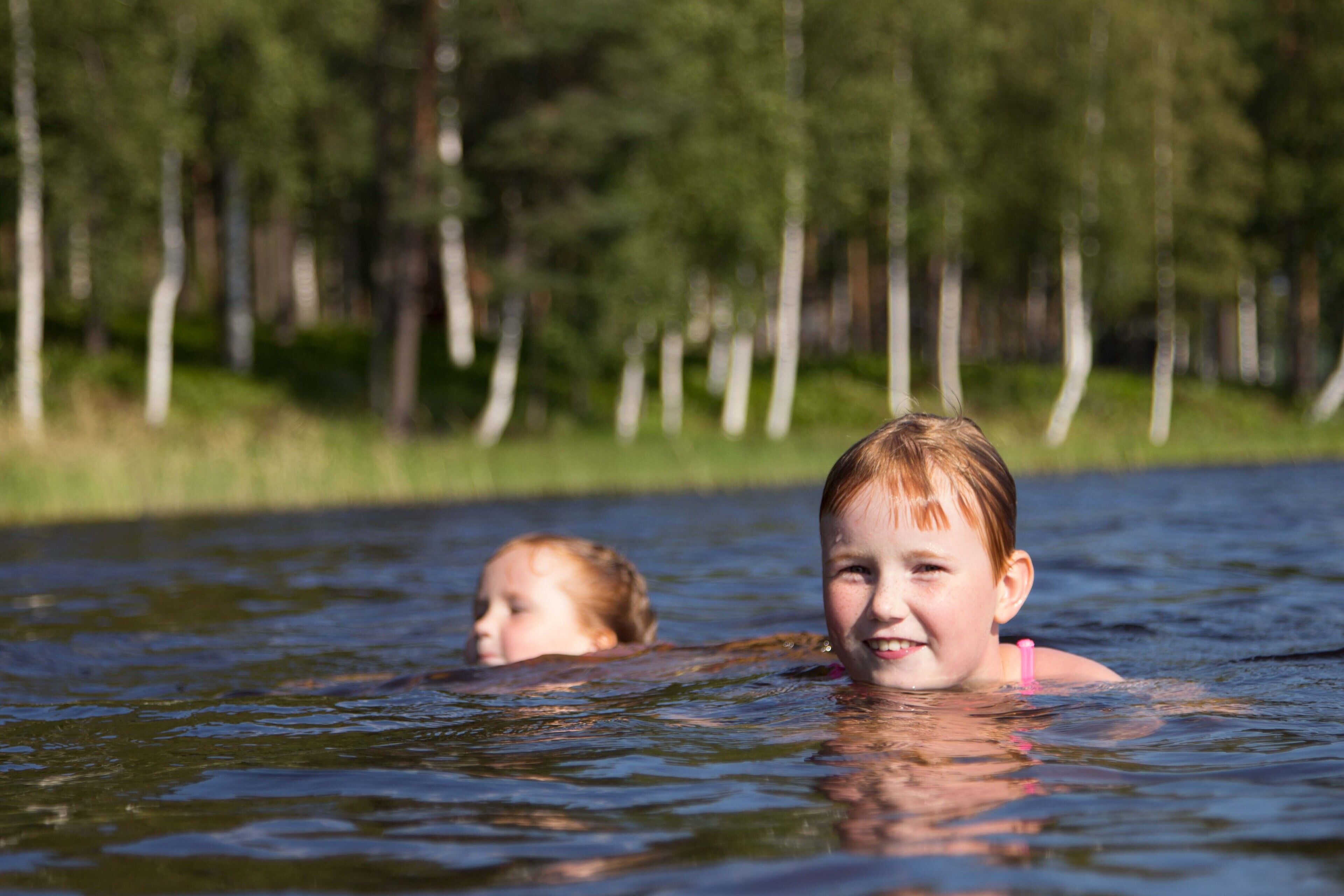 Two children swimming in a body of water with a forested area in the background.