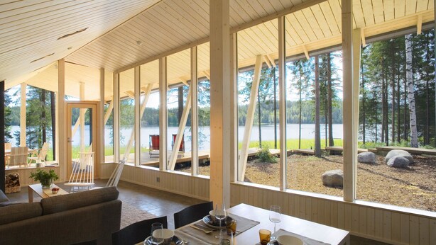 A spacious living area with a large window overlooking a lake, wooden furniture, and a television mounted on the wall. - Anttolanhovi (Mikkeli)