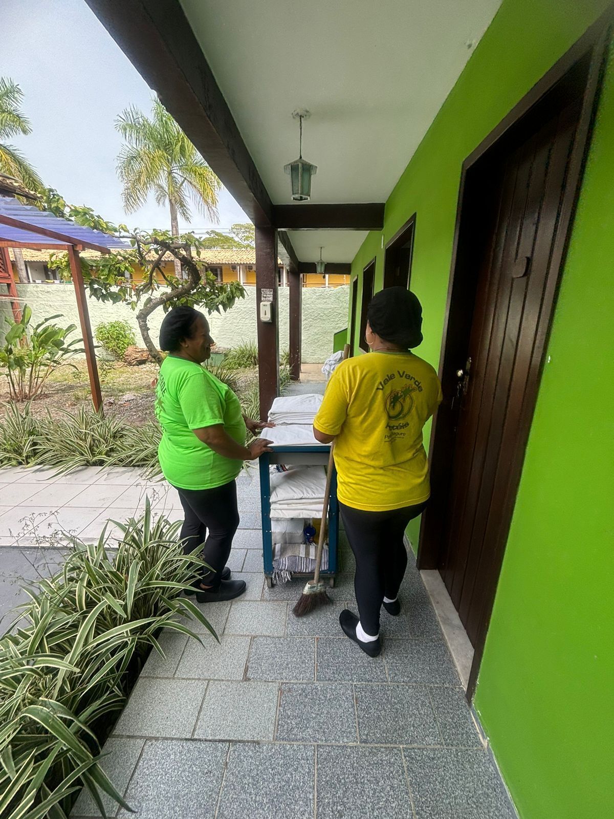 Two individuals outside a building with a green exterior, a covered patio area, and a tiled floor.