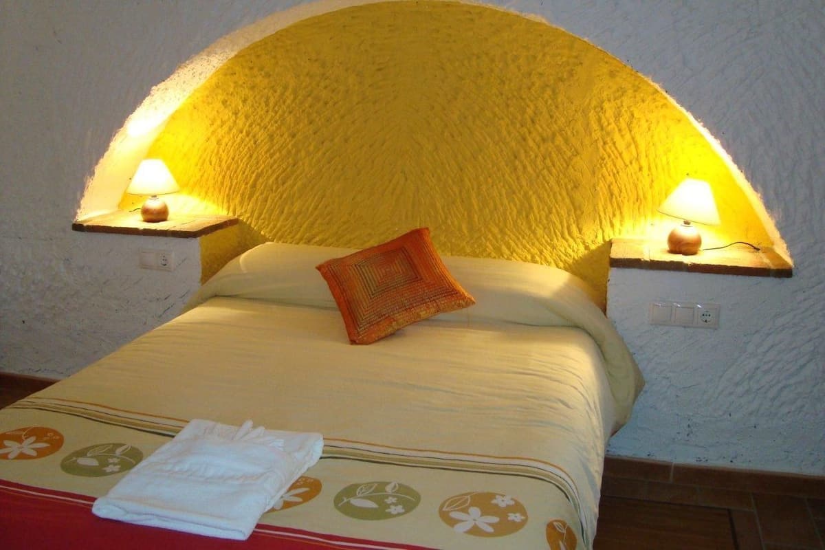 A bed with a yellow textured headboard, two bedside lamps, a red pillow, and a folded white towel on a floral-patterned bedspread.