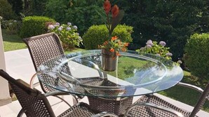 Uma mesa de vidro redonda com cadeiras de vime, uma planta em vaso e uma vista desobstruída de um jardim com sebes e flores.