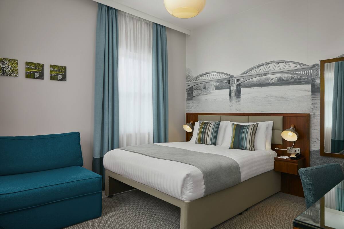 A hotel room with a bed, a blue armchair, a nightstand with a lamp, and a wall mural of a bridge.