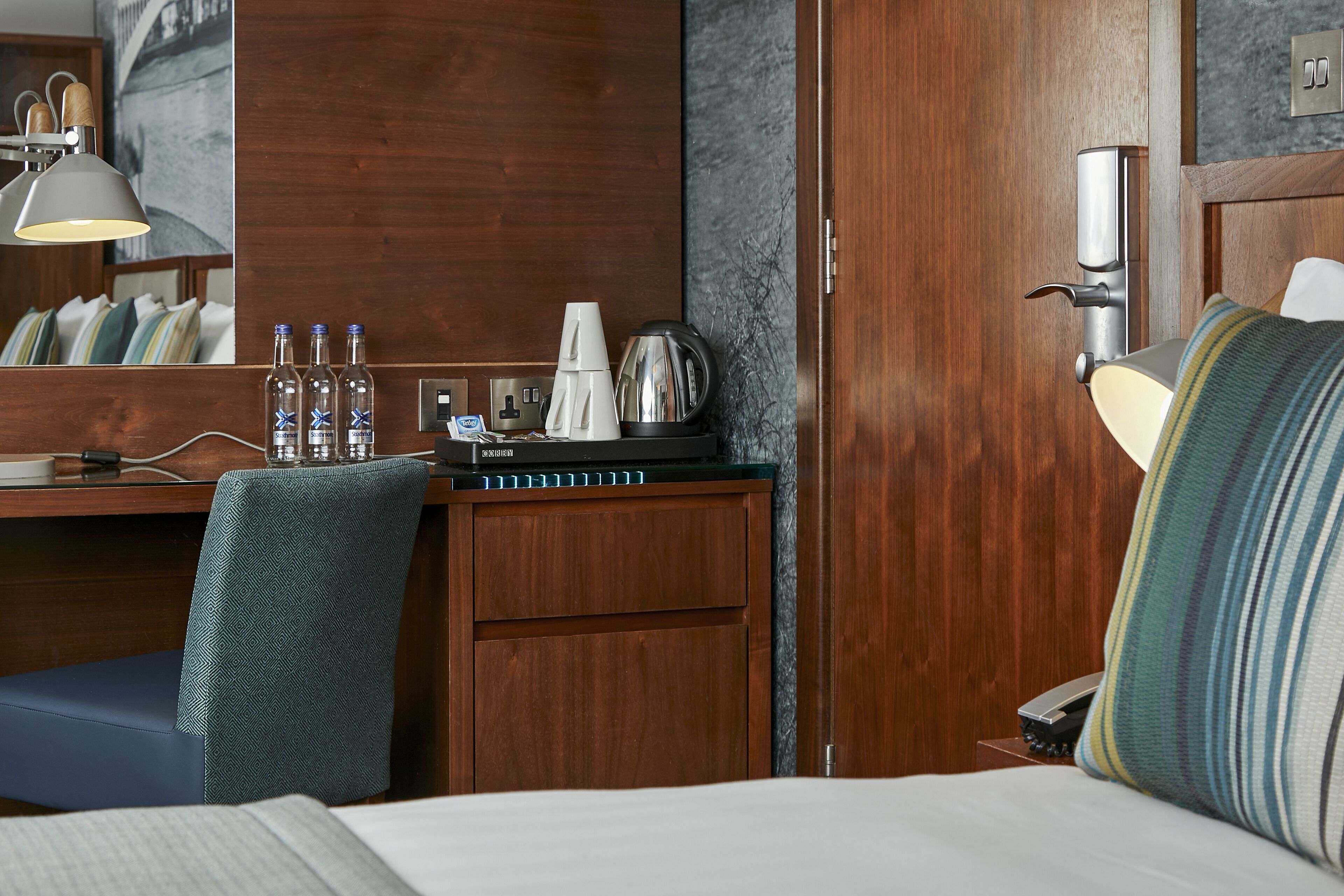 A hotel room with a wooden desk, a chair, a bed, a lamp, and a wall-mounted phone.