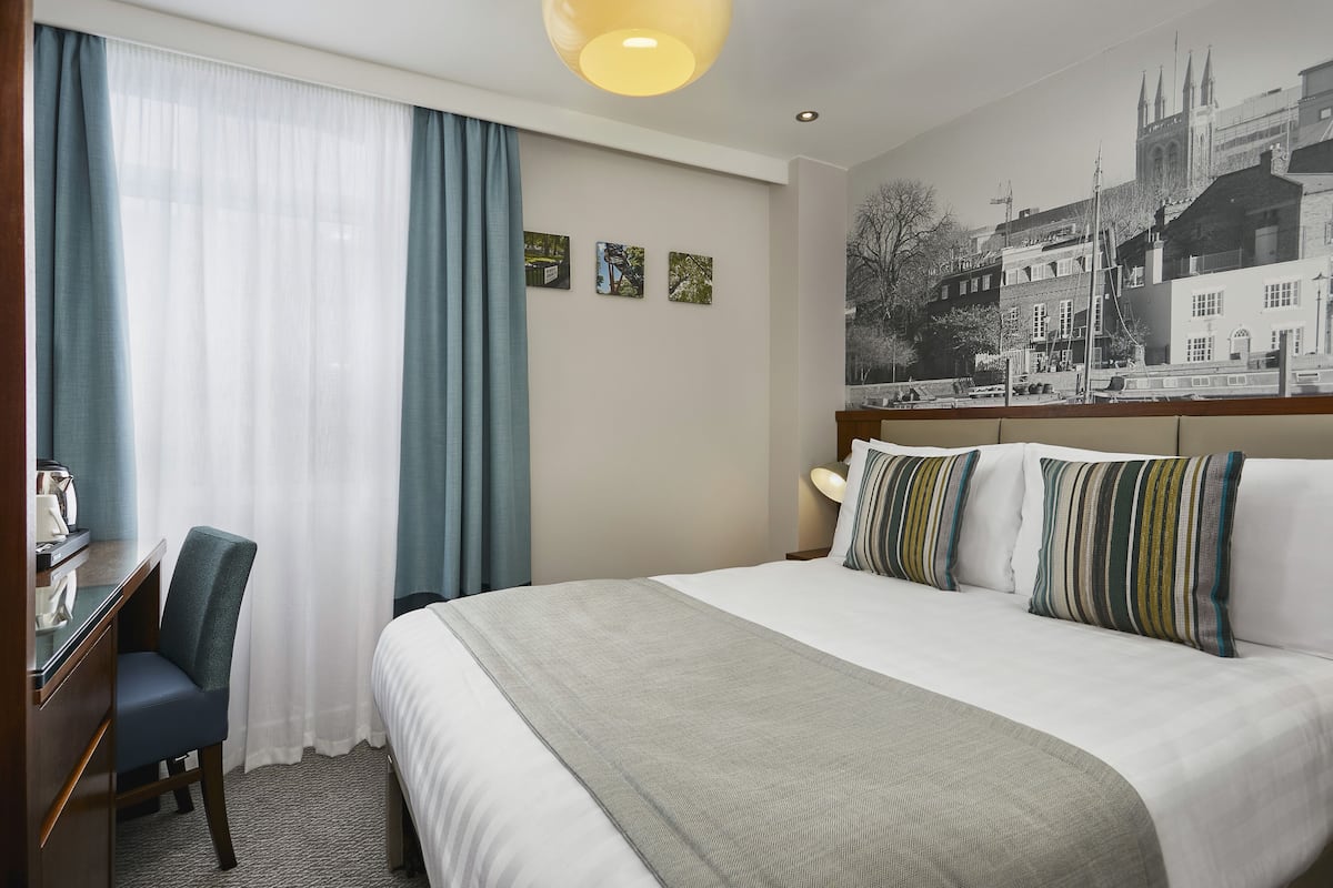 A hotel room with a bed, a desk, a chair, and a large window with curtains. There are three framed pictures on the wall.