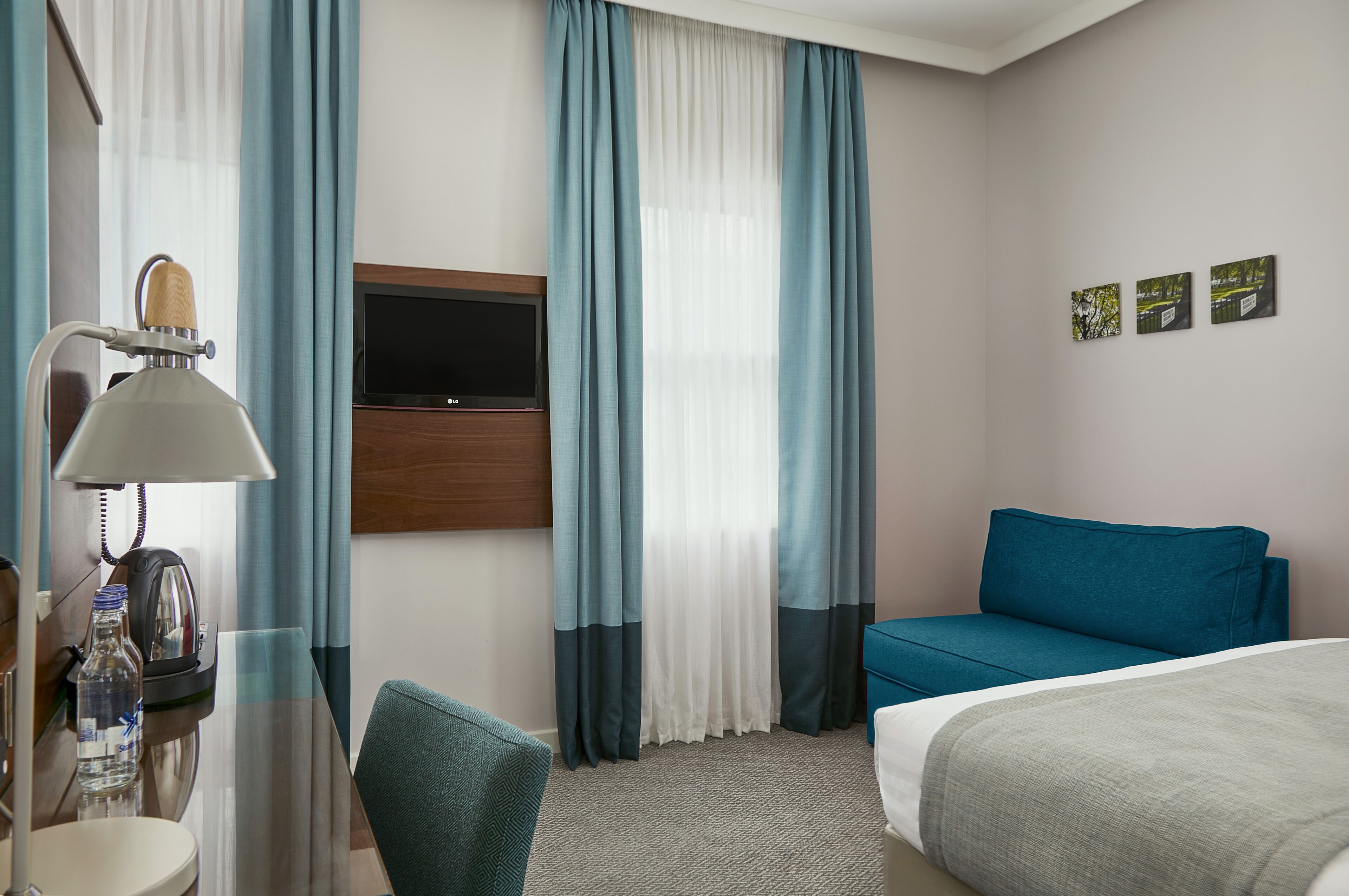 A hotel room with a bed, a sofa, a desk with a lamp, a television, and curtains.