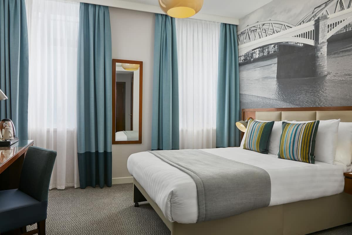 A hotel room with a large bed, a desk with a chair, a mirror, and a view of a bridge through the window.