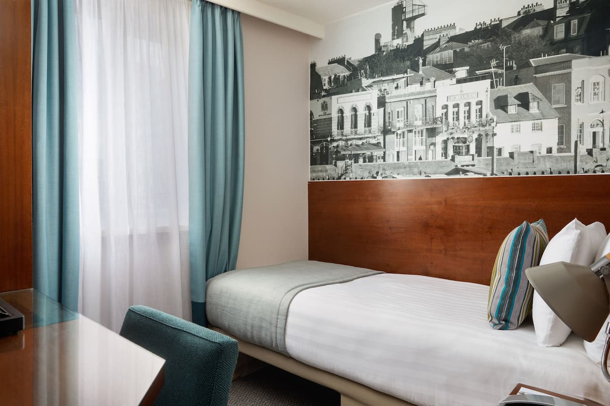 A hotel room with a bed, a wooden headboard, a desk, and a chair. There is a window with curtains, and a black and white cityscape mural on the wall.