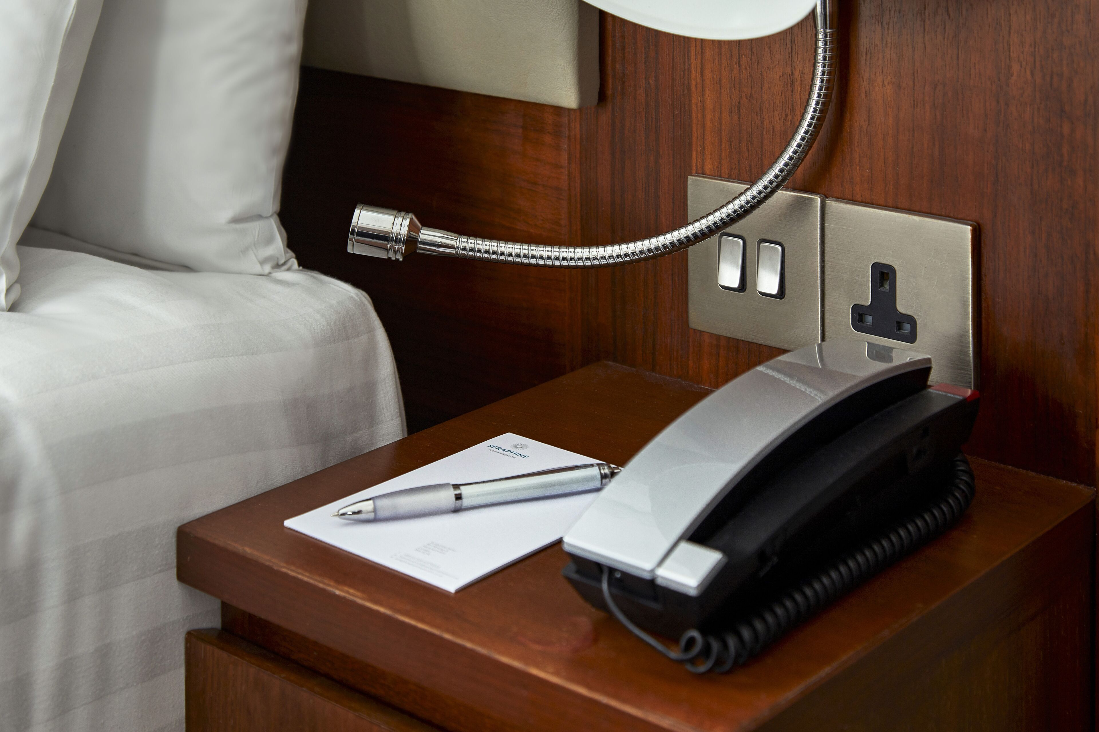 A hotel room with a wooden nightstand, a telephone, a pen, and a document.