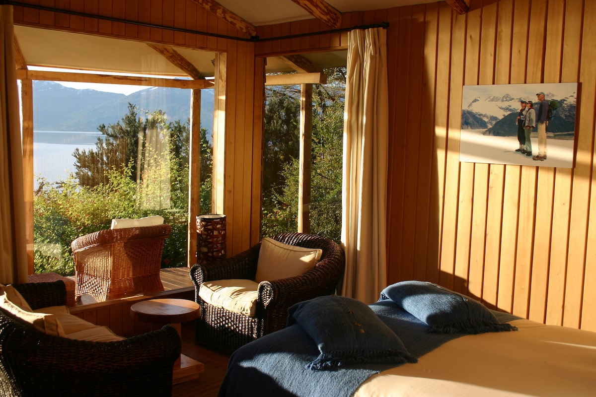 Una habitación con una cama, dos sillones de mimbre, una mesita y un cuadro de un paisaje montañoso.