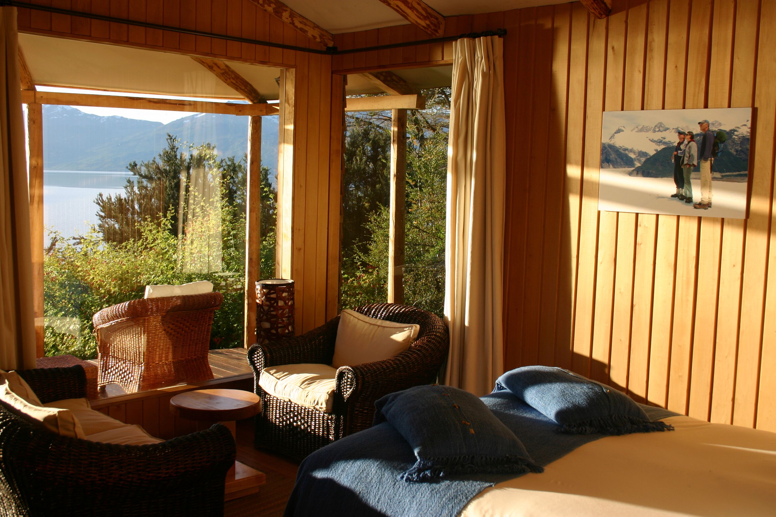 Una habitación con una cama, dos sillones de mimbre, una mesita y un cuadro de un paisaje montañoso.