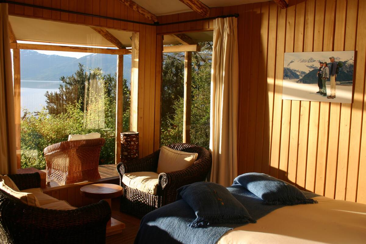 A room with a bed, two wicker chairs, a small table, and a painting of a mountain scene.