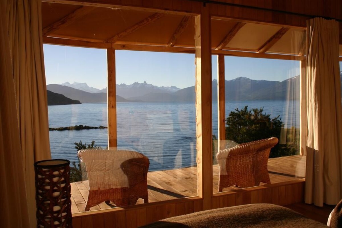 A room with a large window overlooking a lake and mountains, wicker chairs, and a wooden floor.
