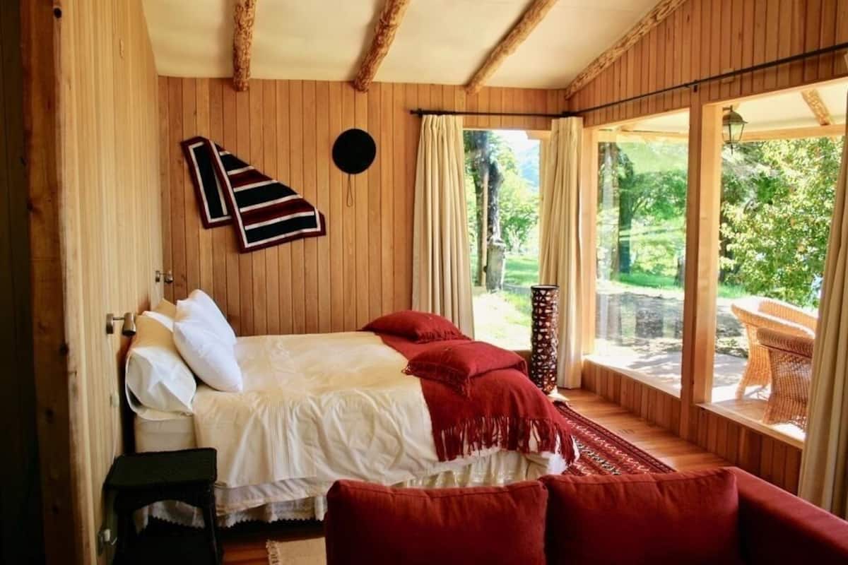 A wooden room with a bed, a small table, and a view of greenery outside.