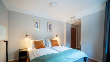 Una habitación de hotel con una cama, dos luces fijadas a la pared, una puerta y un pasillo visible.