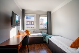 Habitación de hotel con cama, escritorio, silla y televisor. Cuenta con ventanas con cortinas y vistas a edificios.