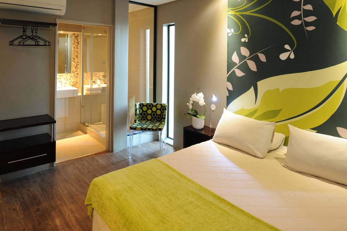 A modern hotel room with a large bed, a bedside table with a lamp, a chair, and a view of the bathroom.