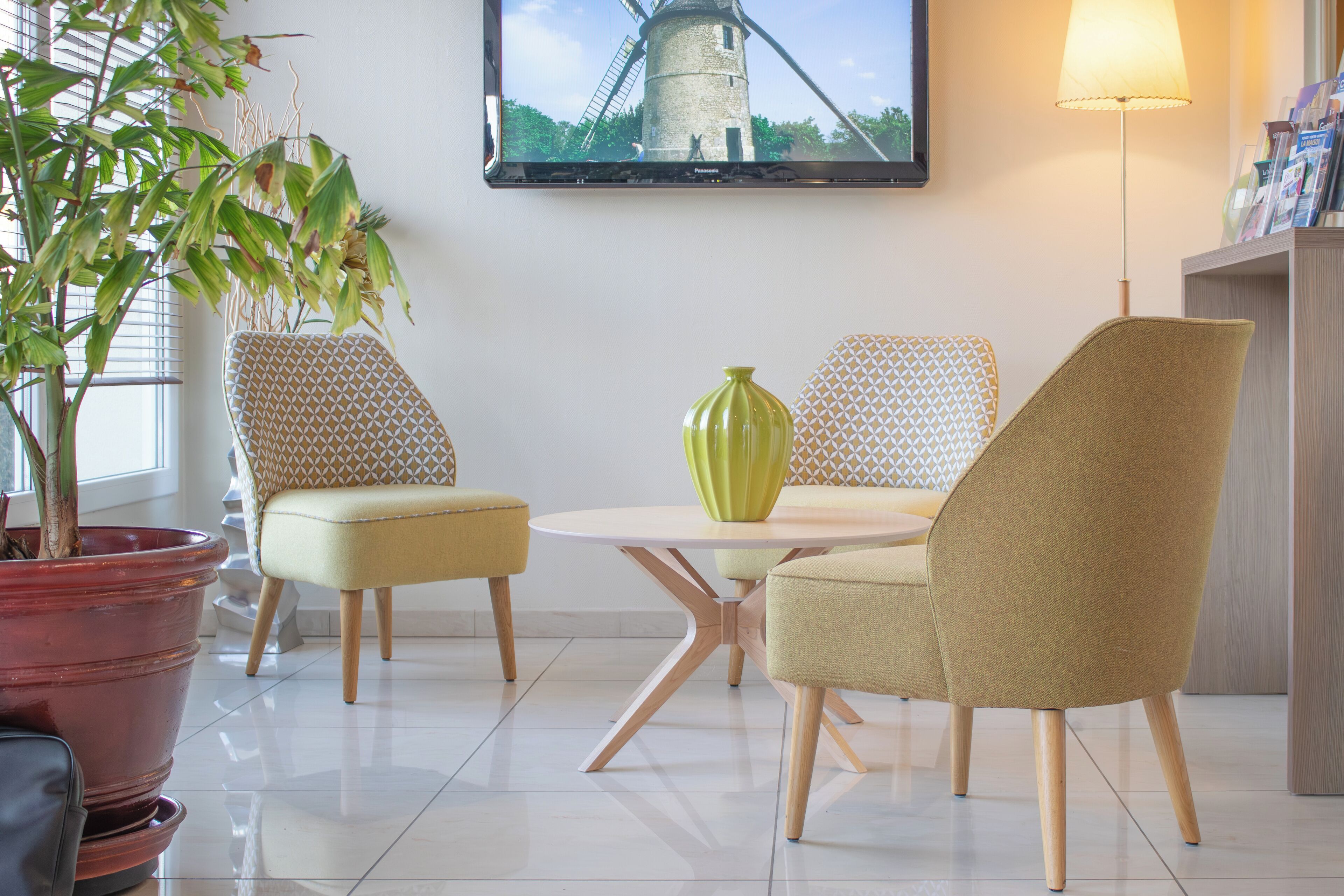Una habitación con una mesa redonda, dos sillas estampadas, un jarrón verde, una planta en maceta y un cuadro en la pared.
