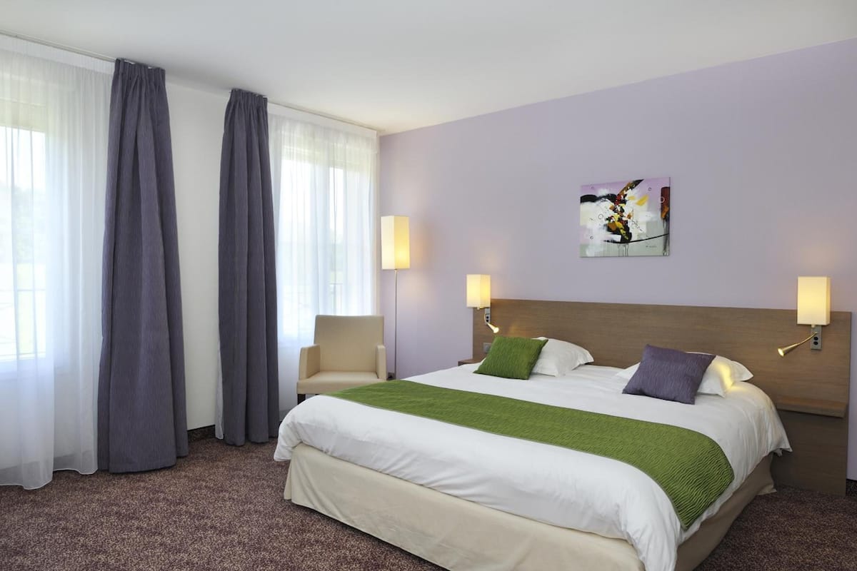 A hotel room with a large bed, two bedside lamps, a green throw blanket, and a painting on the wall.