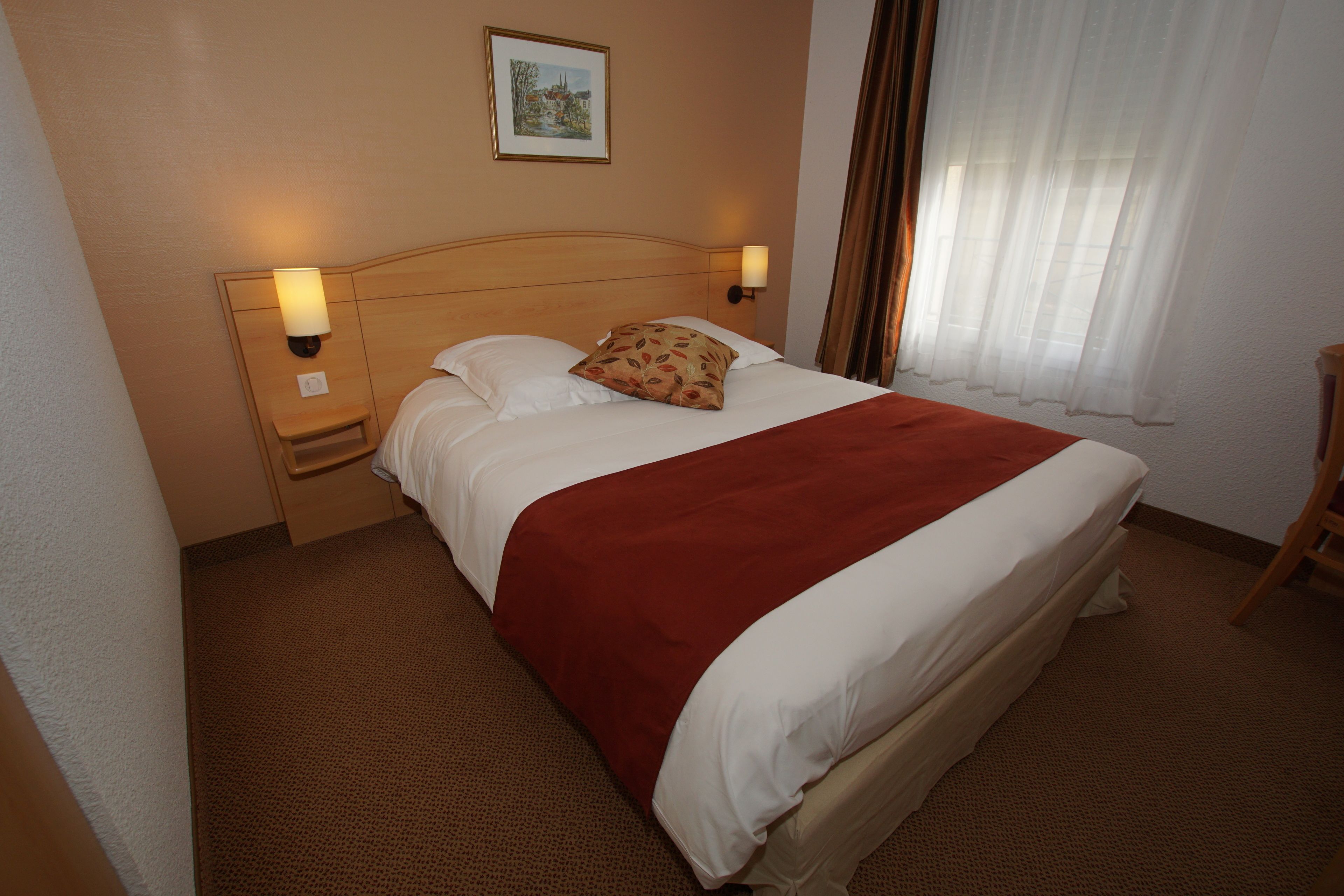 Une chambre d’hôtel avec un grand lit, deux tables de chevet avec des lampes, une tête de lit, un tableau encadré au mur et une fenêtre avec des rideaux.