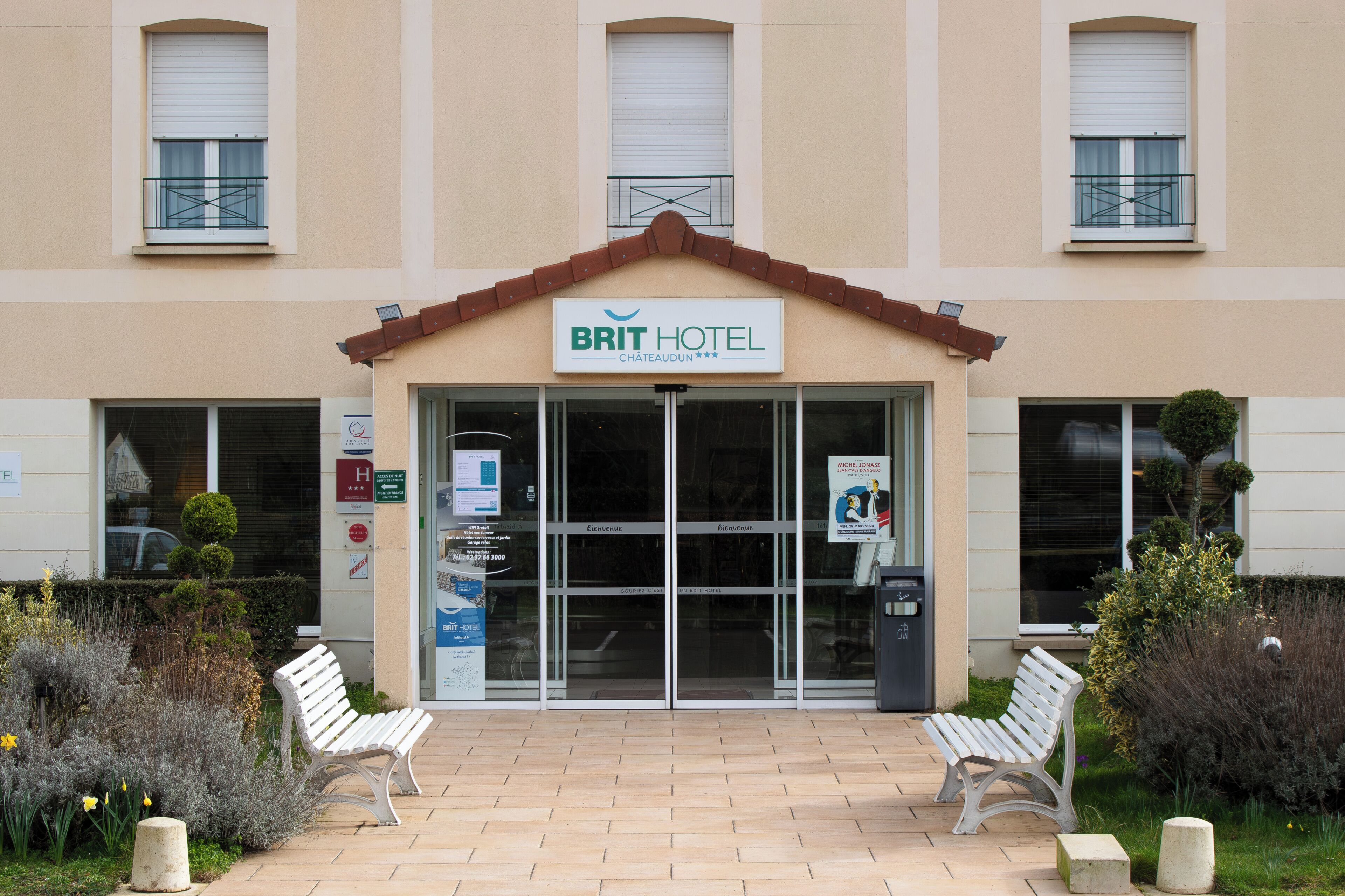 L’entrée d’un hôtel, avec une enseigne indiquant « BRIT HOTEL », deux bancs à l’extérieur et un trottoir dallé.