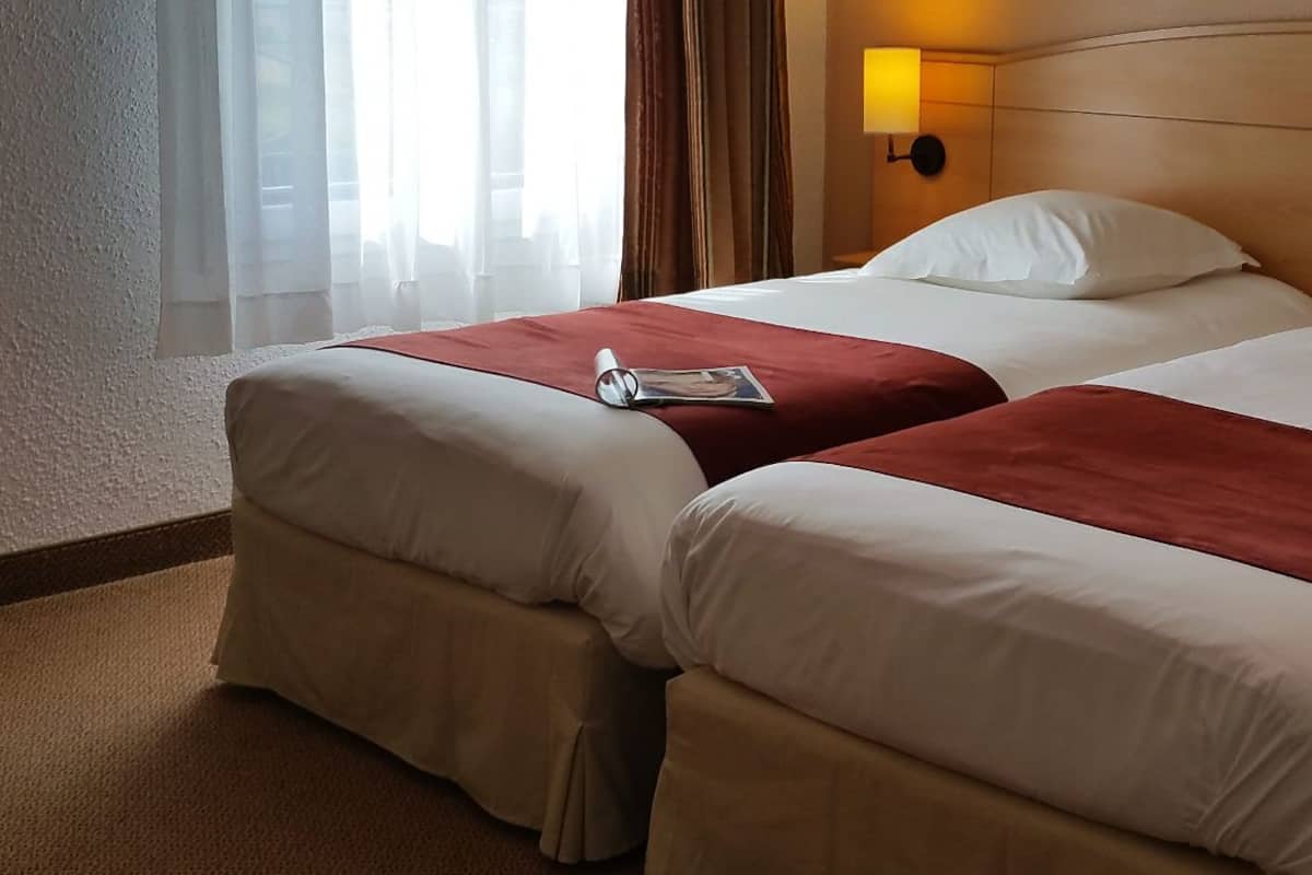 A hotel room with two beds, a nightstand, a lamp, and a window with curtains.
