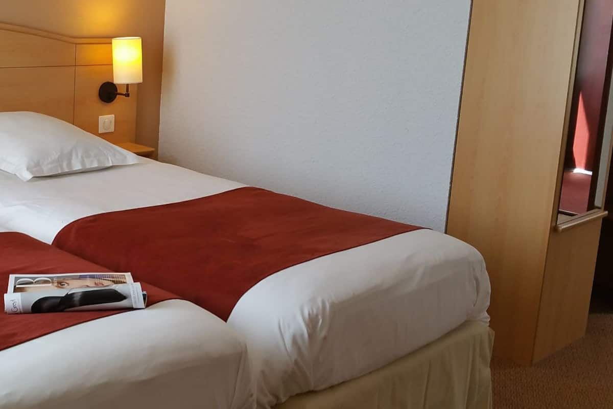 A double bed with white and red bedding, a nightstand with a lamp, and a magazine on the bed.