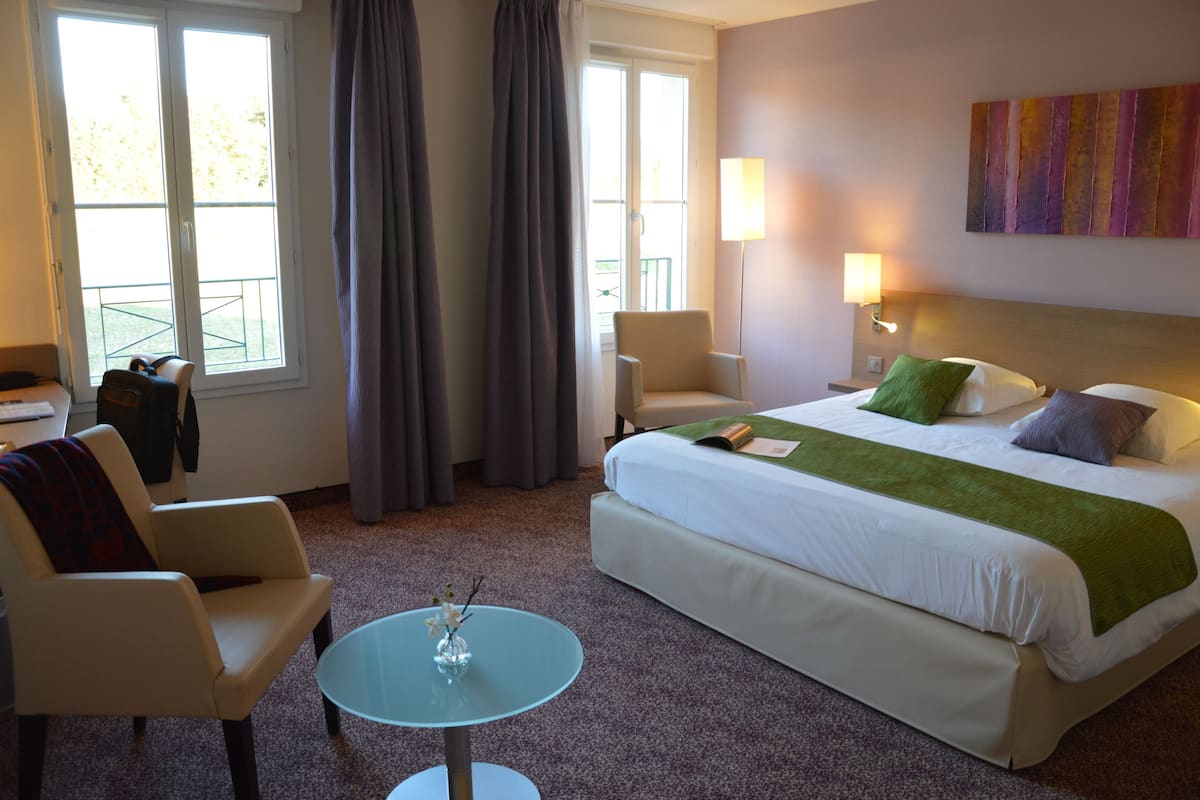 A hotel room with a bed, two chairs, a small table with a vase, a window with curtains, and a painting on the wall.