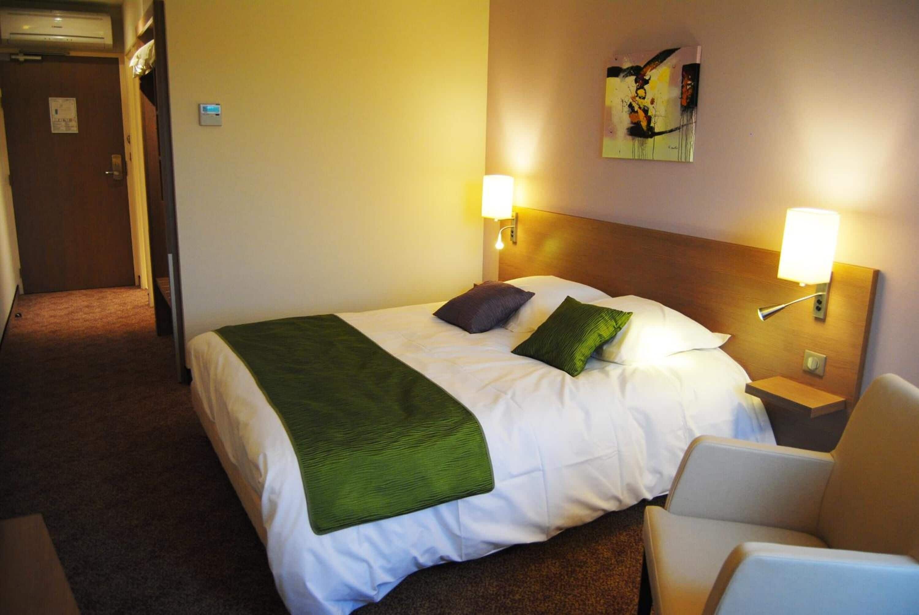 A hotel room with a bed, two bedside lamps, a green throw blanket, a pillow, a headboard, a painting on the wall, and a chair.