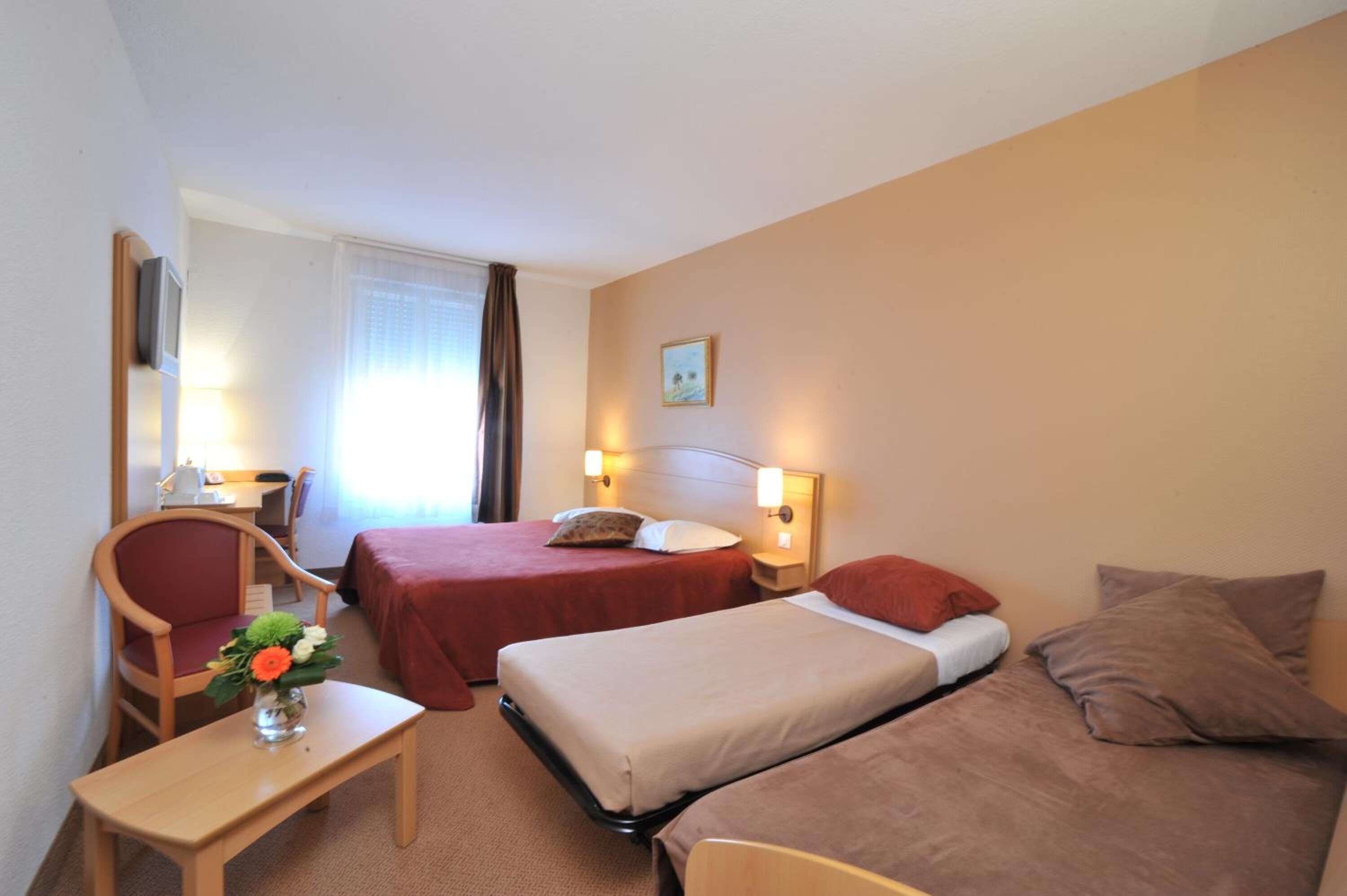 Habitación de hotel con dos camas, un escritorio, una silla y una pequeña mesa con un jarrón de flores.