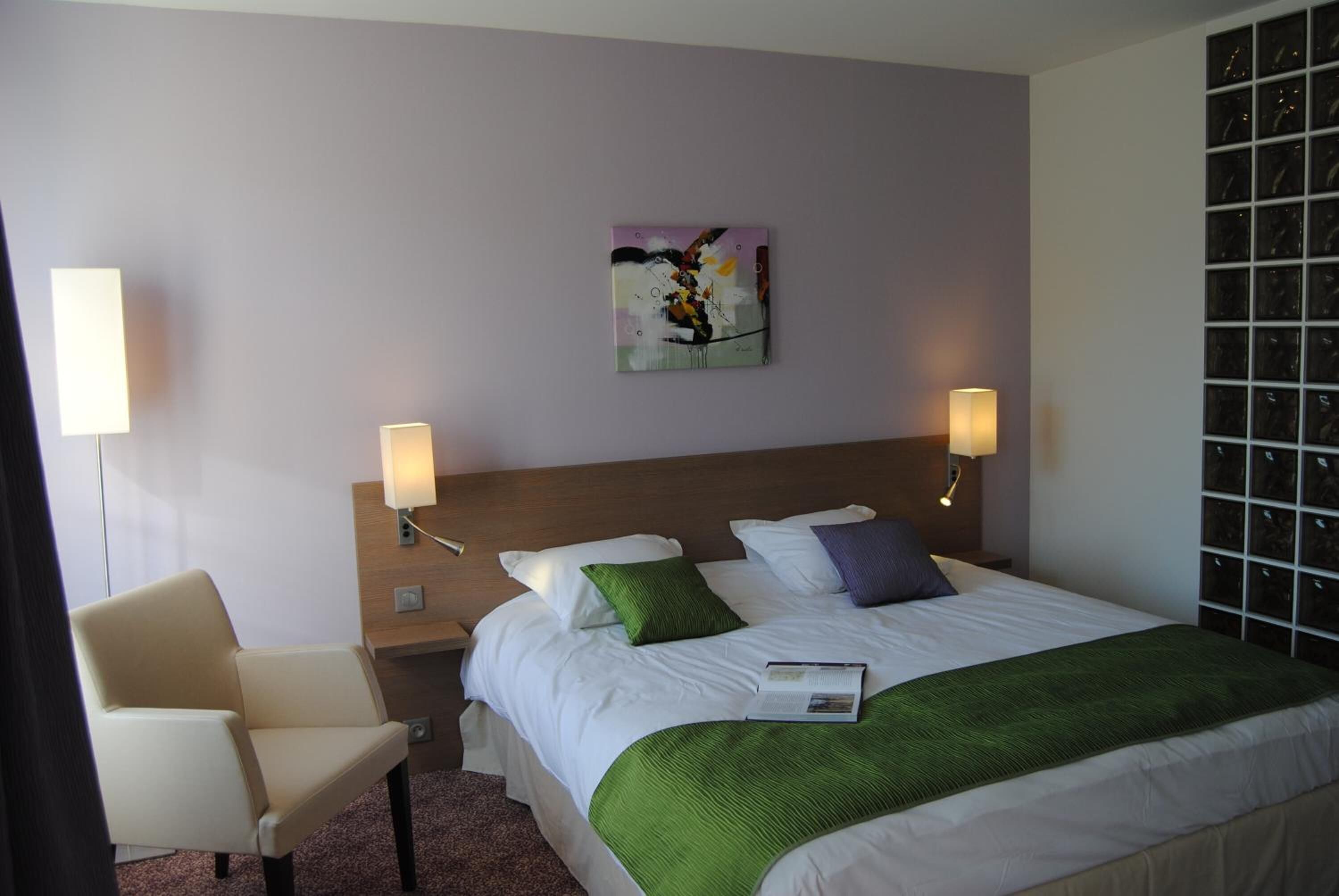 A hotel room with a bed, two bedside lamps, a chair, a painting on the wall, and a window with a view of the city.