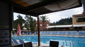 A poolside area with lounge chairs, a table with a menu, and a view of the pool and surrounding buildings. - Christakis Hotel (Corfu)