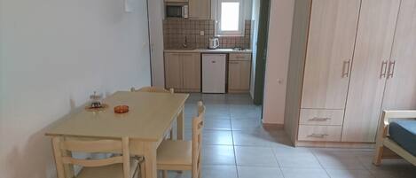 A small, clean kitchen with a microwave, kettle, and refrigerator. There is a dining table with chairs and a cabinet with drawers.