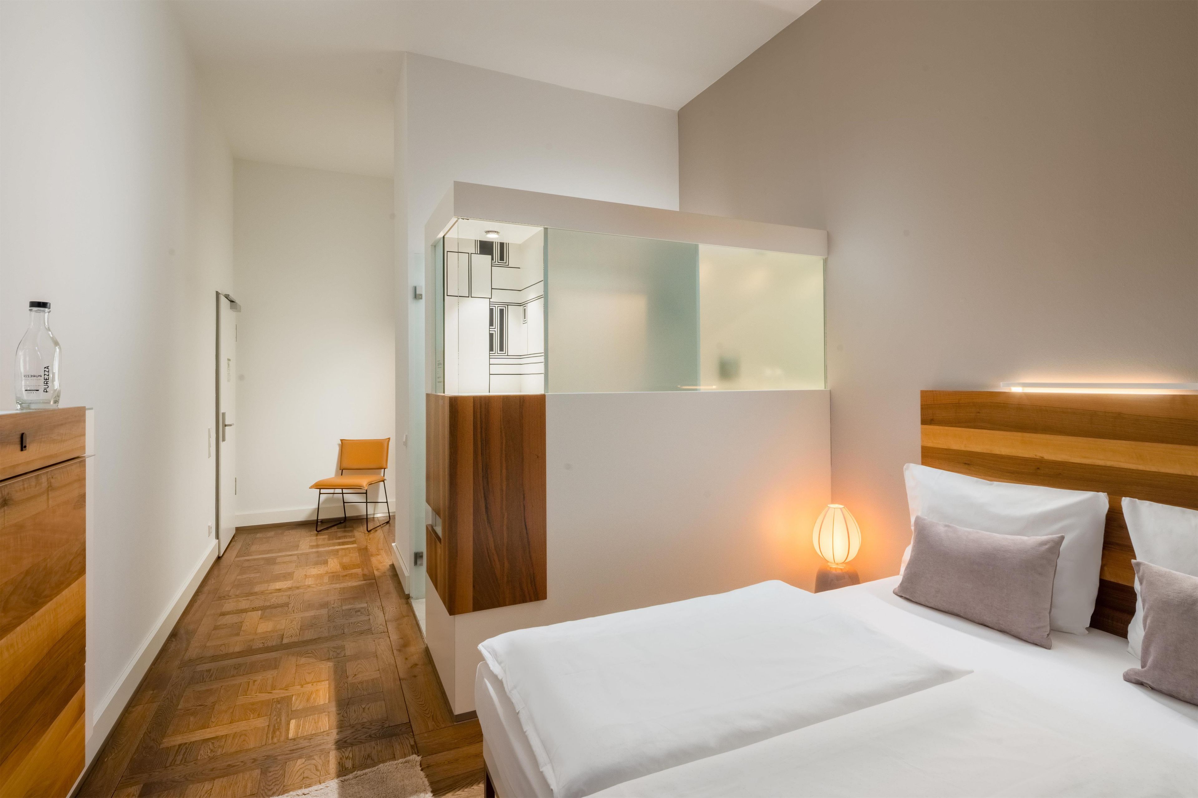 Habitación de hotel con piso de madera, una cama con ropa blanca, una mesita de noche con lámpara y un espejo en la pared.